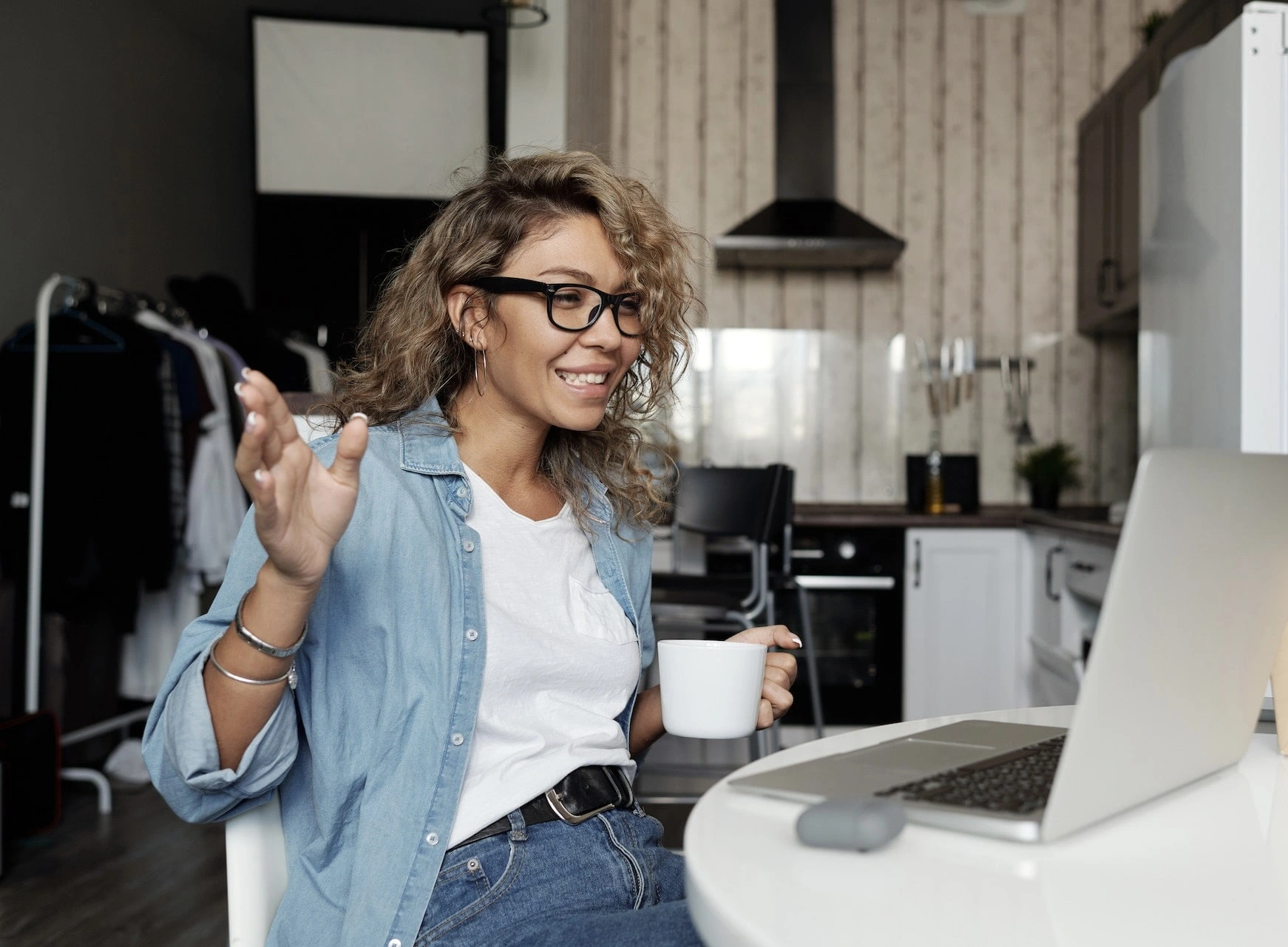 A smiling woman, drinking coffee and talking to someone on her laptop.