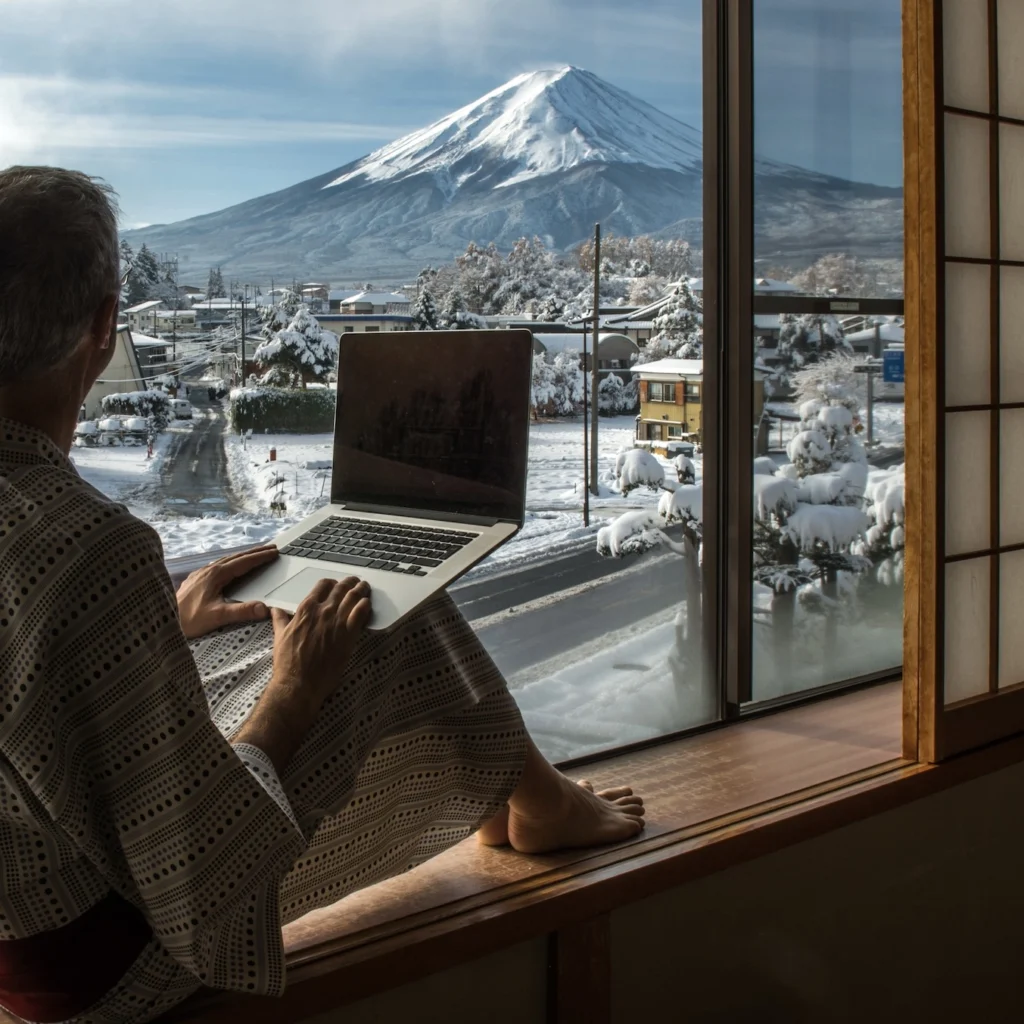A man sitting on windowsill using a laptop while looking at a mountain in the distance. 